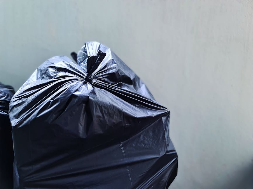 Close-up of a large black plastic rubbish bag, tightly tied at the top, sitting against a plain light-colored wall. The bag appears to be full and slightly crumpled, with visible creases and shiny surface reflecting light. Part of another similar black bag is partially visible to the left, suggesting a collection of waste ready for disposal. The bags are placed on interior flooring or pavement, indicating a setting for waste collection or disposal. The environment appears indoor or within a sheltered area, with minimal background detail. This scene represents the typical appearance of waste prepared for private rubbish removal services, aligning with the context of alternative waste handling methods such as those offered by Rubbish Removal Ilford.