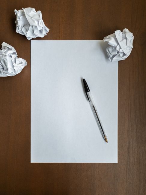 A blank white sheet of paper lies flat on a dark wooden surface, with a black and clear ballpoint pen placed diagonally across it. Surrounding the paper are three crumpled balls of white standard paper, positioned at the top left, top right, and bottom left corners of the image. The lighting appears natural, casting soft shadows around the crumpled paper and the pen, creating a neutral and professional atmosphere. In the background, the wooden surface reflects no significant texture or grain, providing a clean, uncluttered environment suitable for planning or listing tasks. This scene subtly suggests the concept of drafting, editing, or revising written or project ideas, which can relate to organising or preparing for a waste management or rubbish removal service, such as those offered by Rubbish Removal Ilford, especially in contexts involving on-site planning or alternative waste handling arrangements.