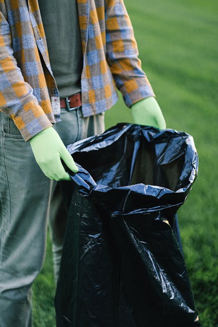 A person wearing a plaid shirt with shades of yellow, blue, and gray, alongside a green long-sleeve shirt and beige trousers secured with a brown belt, is holding open a large black rubbish bag lined with a shiny, metallic interior. The individual is standing outdoors on a lush green lawn, with their hands gloved in bright green disposable gloves. The rubbish bag is partially filled and positioned vertically, ready for waste collection or disposal. The background features a well-kept grassy area, suggesting a garden or park environment. The scene depicts an act of waste collection or cleanup, aligning with private or independent rubbish removal activities often handled by services like Rubbish Removal Ilford, particularly relevant within the context of on-site clearance and alternative waste handling methods typically used when conventional local authority collection is unavailable or delayed.