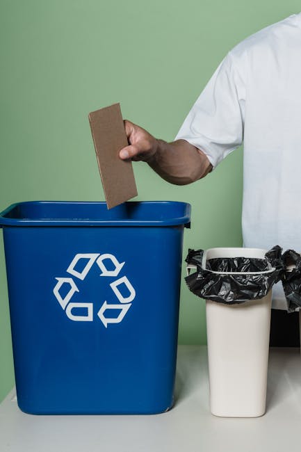 A person wearing a white shirt is holding a rectangular piece of cardboard in their right hand, positioned above a blue recycling bin with a white recycling symbol on the front. The bin is placed on a light gray surface against a plain, light green wall background. To the right of the blue bin, there is a smaller, off-white waste container lined with a black plastic bag, which appears to be used for general rubbish. The scene is indoors, with natural or neutral lighting illuminating the objects and emphasizing their textures and colors, illustrating an aspect of waste separation or on-site rubbish handling that could be associated with private disposal services such as those offered by Rubbish Removal Ilford, especially within a context of managing waste in flats or for landlords.