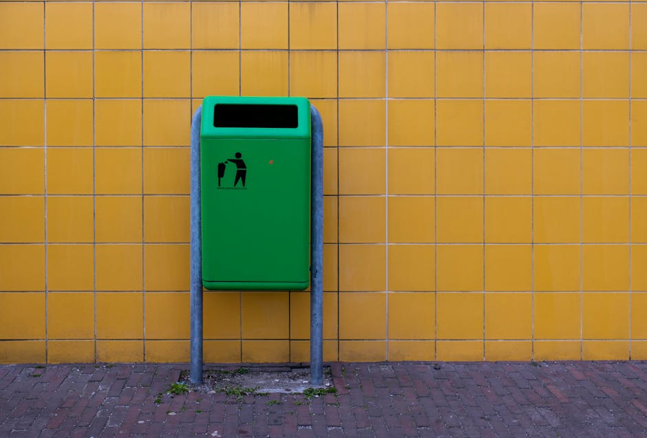 A collection of overflowing waste and recycling bins situated on a paved area near parked cars, with various black, red, and grey containers among large piles of mixed rubbish including cardboard boxes, paper, plastic bags, and discarded packaging. The scene is set against a background of a commercial building with storefronts, some with closed shutters, and a blue metal scaffolding structure above. The waste is spilling onto the pavement, with some items on the curb and ground, indicating an unmanaged rubbish collection situation often associated with private disposal services rather than standard council collections. A small tree is visible on the left side, with a metal railing separating the waste area from the parked vehicle and pedestrian pathway. The overall environment suggests an urban setting where waste management may require additional attention from local rubbish removal services like Rubbish Removal Ilford, highlighting the importance of proper waste disposal rules for flats and landlords.