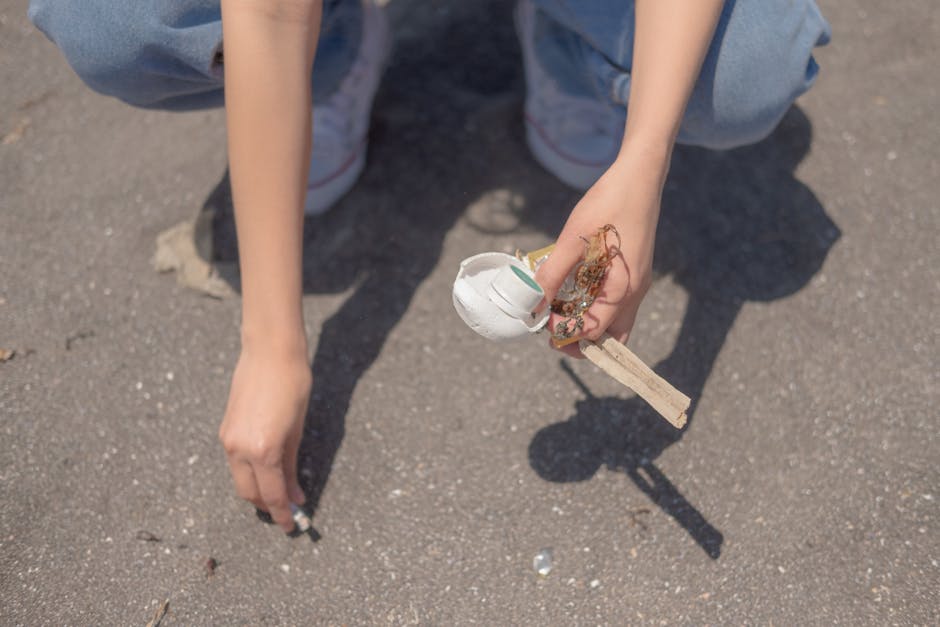 A person wearing light blue jeans and white sneakers is crouched on a paved surface, holding a small, broken ceramic or plaster object with visible jagged edges and a rough, uneven surface. The object appears to be a fragment of a larger piece, with some dirt and possibly plant material attached. The person's right hand gently grips the fragment, while their left hand is near the ground, possibly supporting or steadying themselves. The background shows the textured asphalt pavement, with no other objects or environmental features clearly visible. The lighting appears natural, suggesting an outdoor setting. This scene reflects a context of tidying or clearing debris, which correlates with waste removal or rubbish collection activities, such as those offered by Rubbish Removal Ilford.