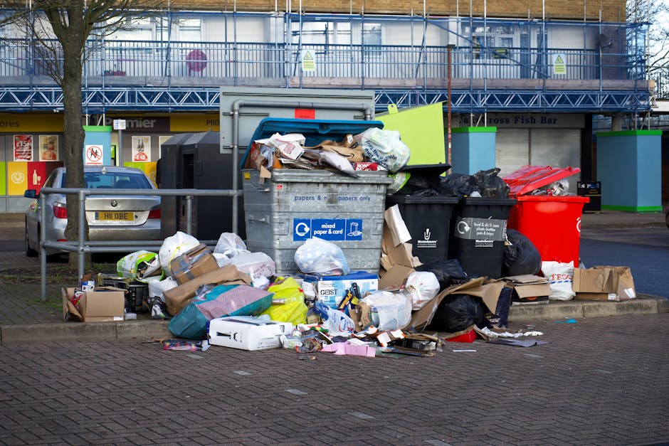 A large collection of overflowing rubbish bins and scattered waste, including cardboard boxes, plastic bags, paper, and packaging materials, is piled on a paved area adjacent to a parking space. The waste appears to consist of mixed household and commercial refuse, with some items spilling onto the surrounding pavement. Behind the disposal units, there are parked cars and a multi-storey building with a blue and green storefront on the ground level, indicating a commercial area. The bins include a gray mixed paper and card bin, black general waste bins, and a red recycling container, all situated near metal railings that separate the waste area from parked vehicles. The scene is captured under natural daylight, with a tree visible on the left side and an orange and green exterior decor around the building, suggesting an urban or suburban environment. The image depicts an instance where independent rubbish collection or private waste handling by services like Rubbish Removal Ilford may be necessary to manage overflowing refuse on a commercial or residential frontage.