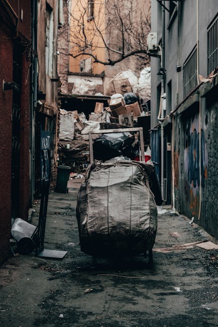 A narrow urban alleyway filled with various types of waste and discarded materials, mainly consisting of large black and white garbage bags, some torn or partially collapsed, with a wheeled trolley or cart in the foreground used for rubbish collection. Behind the trolley, there are piles of cardboard boxes, plastic bags, and other refuse stacked against a wooden fence and a brick wall, which are weathered and stained from exposure. To the right, a building with metal grilles on the windows and grey painted exterior walls is visible, while the left side features graffiti artwork on the wall. In the background, leafless tree branches are visible above the waste heaps, with a muted, overcast sky contributing to a subdued and slightly neglected atmosphere typical of an area where private rubbish collection or waste removal services like Rubbish Removal Ilford might be engaged for local junk clearance or alternative waste handling. The environment appears damp, with dark patches on the asphalt ground, reflecting a cold or rainy day, emphasizing the need for proper waste disposal solutions.