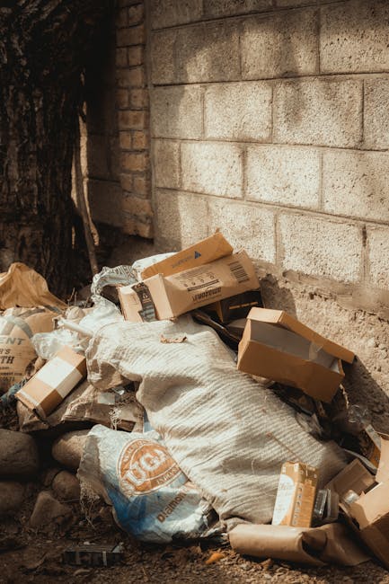A pile of discarded cardboard boxes and paper packaging waste, some flattened and some partially crumpled, resting against a rough-textured brick wall and a large, dark tree trunk in an outdoor setting. The boxes are in various shades of brown, with printed labels and barcodes visible on some, and are mixed with woven fabric sacks or bags that appear coarse and slightly dirty. The scene is illuminated by natural daylight, casting soft shadows on the ground, which consists of dirt, small stones, and debris. The background includes the brick wall and the tree, indicating a setting near a property boundary or alleyway, suggesting an instance of private rubbish disposal or clearance possibly managed by a waste removal service like Rubbish Removal Ilford. The overall composition reflects a typical scene of unwanted waste ready for collection or removal, aligned with professional rubbish management practices.