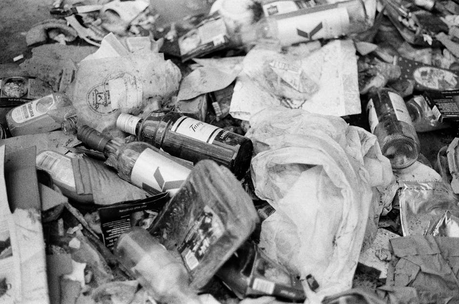 A large pile of mixed waste materials scattered across an outdoor surface, including crumpled paper, plastic bottles, cardboard packaging, and tin cans. The waste appears discarded and not sorted, with some items partially compressed or torn. Visible on the ground are plastic and paper waste, with some transparency or glossy finishes on packaging materials. The bottles are mainly transparent with labels still attached, and the cardboard appears flattened and soiled. The background shows additional waste and debris, creating a dense, cluttered environment typical of uncontrolled rubbish accumulation. The lighting is natural, with shadows indicating outdoor conditions. This scene exemplifies the type of rubbish that a professional waste removal service like Rubbish Removal Ilford may handle through on-site clearance or alternative waste handling methods, emphasizing the importance of proper disposal and recycling of household or commercial waste.