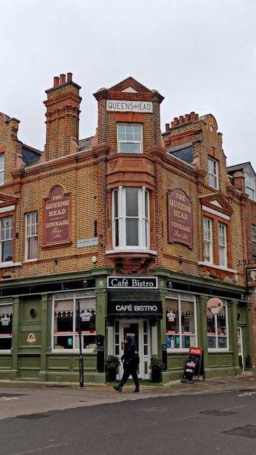 A street scene on Ilford High Road featuring a mix of historic and modern brick buildings with detailed architectural facades, including arched windows, decorative cornices, and contrasting white trim. The ground floor of several buildings hosts small commercial establishments, such as a restaurant named Bella Italia with blue signage and awnings, and other retail units. In the foreground, a paved roadway with visible double yellow lines runs across the bottom of the image, and a small area of sidewalk is visible with pedestrians walking along the street. Two double-decker buses are parked adjacent to the curb, partially obscuring some storefronts, and various pedestrians are engaged in their activities, some carrying shopping bags, while others are strolling or waiting to cross. The environment appears to be a busy, well-maintained town centre with natural daylight casting soft shadows, indicative of late afternoon or early evening, providing a neutral, professional, and observational depiction suitable for content related to local rubbish removal or private waste disposal services by Rubbish Removal Ilford.