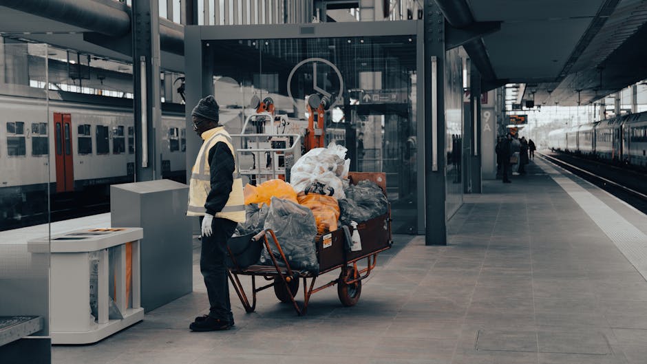 A person dressed in dark clothing and a grey beanie hat stands on a train station platform next to a large metal trolley cart filled with various types of waste bags, including black, grey, and orange plastic, as well as some larger white bags. The trolley is positioned near a glass shelter with a door, behind which a railway line with a train is visible, along with a few other passengers walking in the background. The platform is made of dark, smooth paving stones, and the station features modern architecture with metal supports and a partially covered roof. The environment is lit with natural daylight, creating a clear and neutral setting suitable for discussing private waste handling or alternative rubbish collection services, as seen in the presence of waste accumulation ready for disposal or collection by an independent rubbish removal service such as Rubbish Removal Ilford.