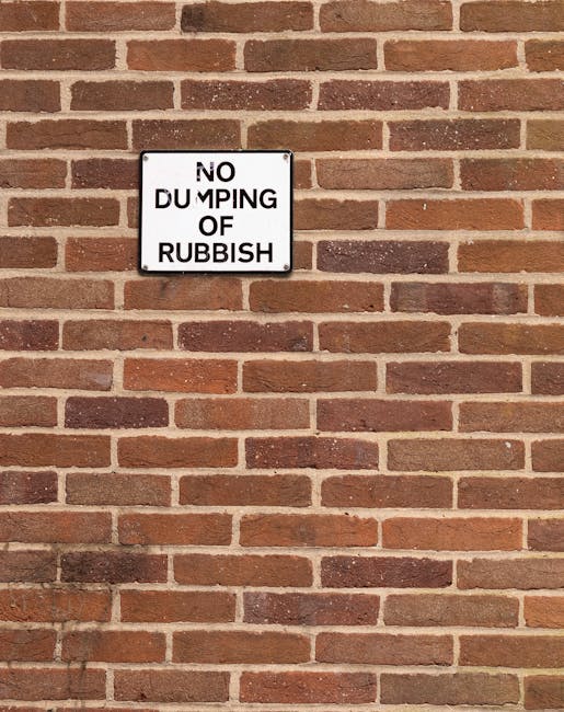 A red metal rubbish skip with visible rust and a weathered surface is positioned on a paved area against a modern dark green and grey wall of a building. Next to the skip, several white plastic rubbish bags filled with waste are stacked at the base, appearing loosely tied. The skip is situated near the corner where the green wall meets a light grey concrete wall, with a small, dark window visible on the building's façade above. The environment appears to be an urban outdoor space, possibly an alley or side access route, with ambient daylight illuminating the scene. The setting exemplifies a typical private rubbish collection or on-site waste clearance by a service such as Rubbish Removal Ilford, highlighting the type of waste management occurring outside commercial or residential premises, where waste is staged for disposal or collection. The overall scene captures an efficient example of independent waste handling in an urban context, with the focus on the debris ready for removal, aligned with professional rubbish removal services.