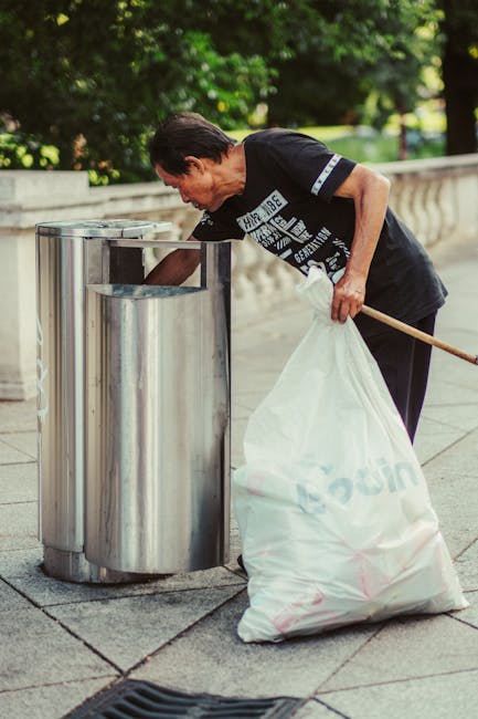 A middle-aged man with dark hair, wearing a black t-shirt with white text on the front, is seen disposing of rubbish into a tall, cylindrical stainless steel bin located outdoors on a paved surface. The man is slightly bent forward, holding a wooden stick in his right hand and grasping a large white garbage bag filled with waste in his left hand. The bin has a shiny, reflective surface and is situated against a low stone wall that runs along a park or garden area. Behind him, there are lush green trees and foliage, providing a natural backdrop under daylight conditions. The scene appears to be part of a public space, emphasizing the importance of waste disposal and cleaner environments, which aligns with private rubbish collection services like those offered by Rubbish Removal Ilford in the context of alternative waste management and rubbish removal solutions.